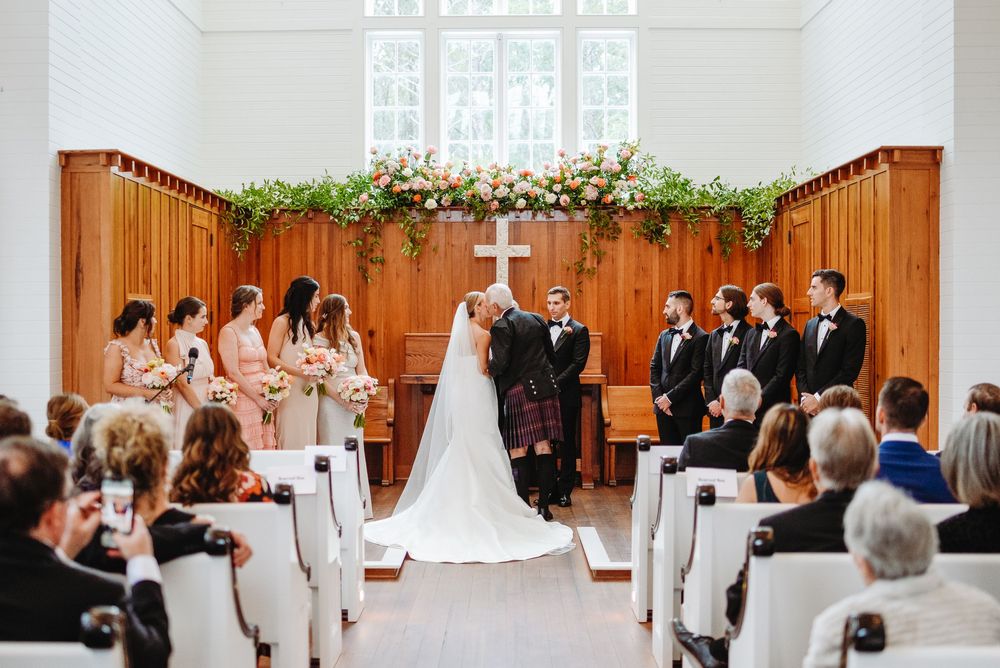 Wedding ceremony in a white chapel with wooden accents and greenery garlands decorating the altar area.