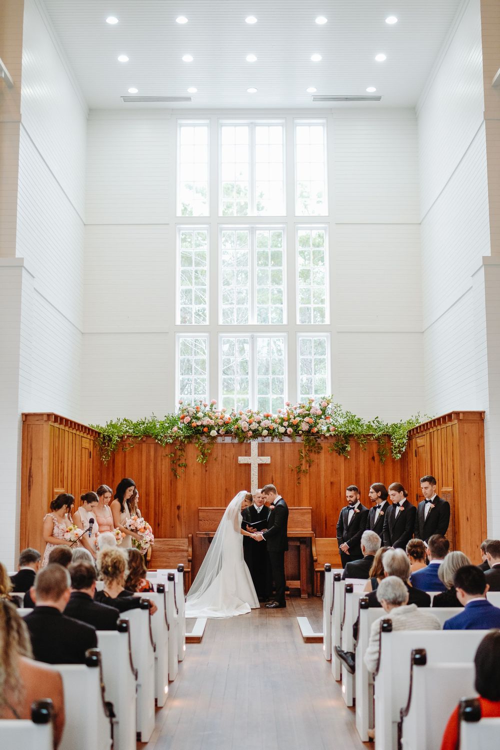 Elegant wedding ceremony taking place in a bright chapel with white walls, wooden paneling and guests seated in white chairs.