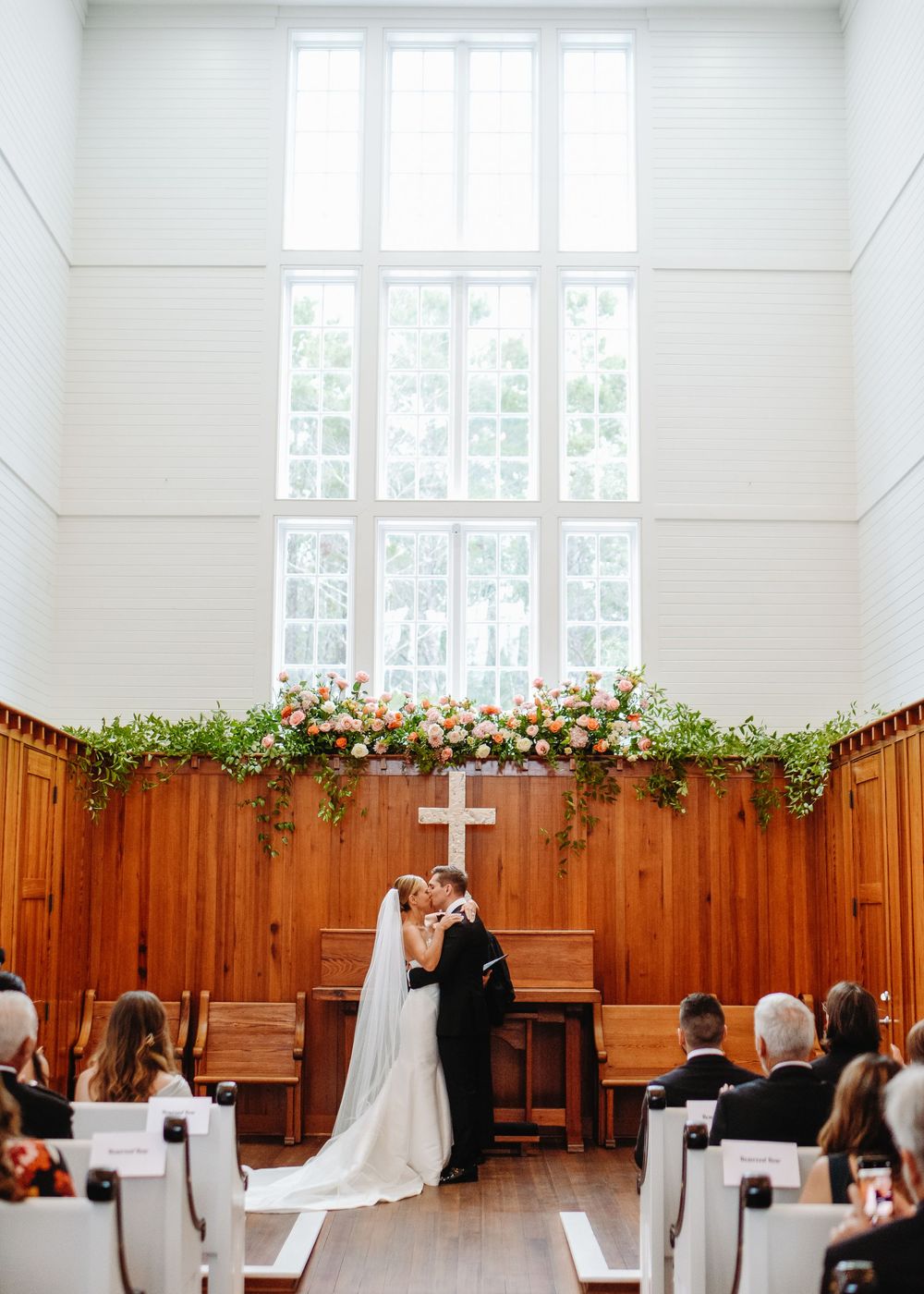Elegant church wedding ceremony with wooden paneled walls decorated with floral arrangements and a white cross.