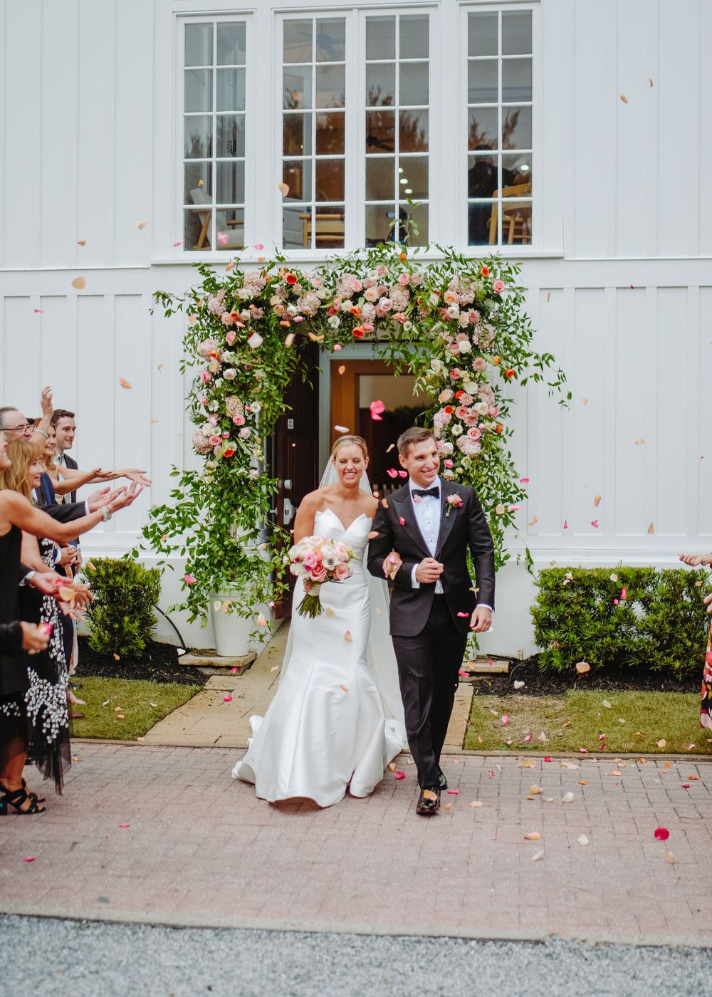 Wedding couple and guests exit venue while flower petals are tossed in celebration along paved path.