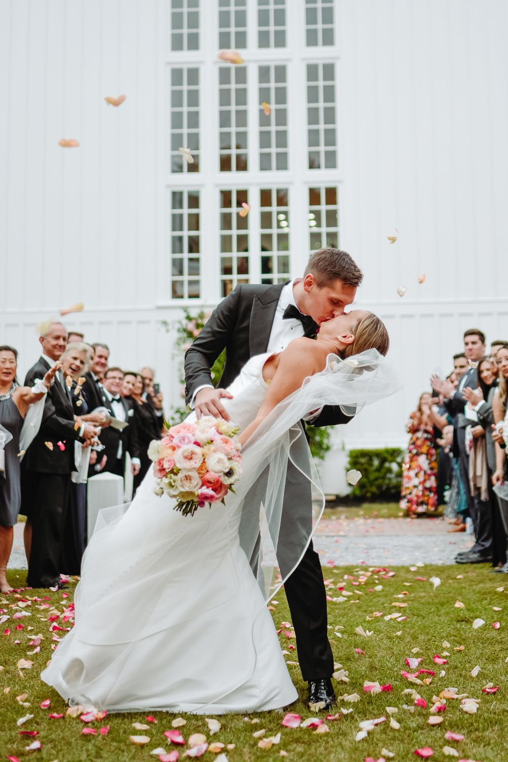 Romantic wedding couple performs traditional dip pose during outdoor celebration as guests throw rose petals.