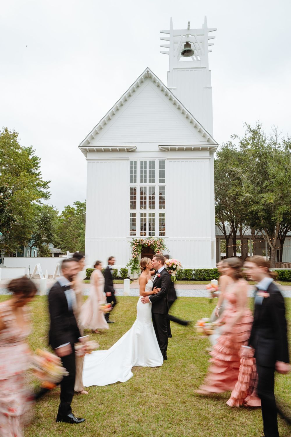 Motion-blurred wedding party dancing in front of a white chapel building.