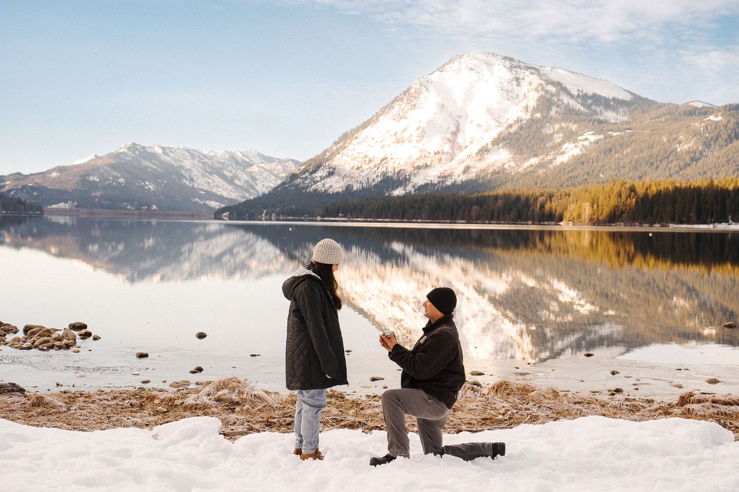 Two people enjoying a winter view of a snow-capped mountain reflecting perfectly in a calm lake.