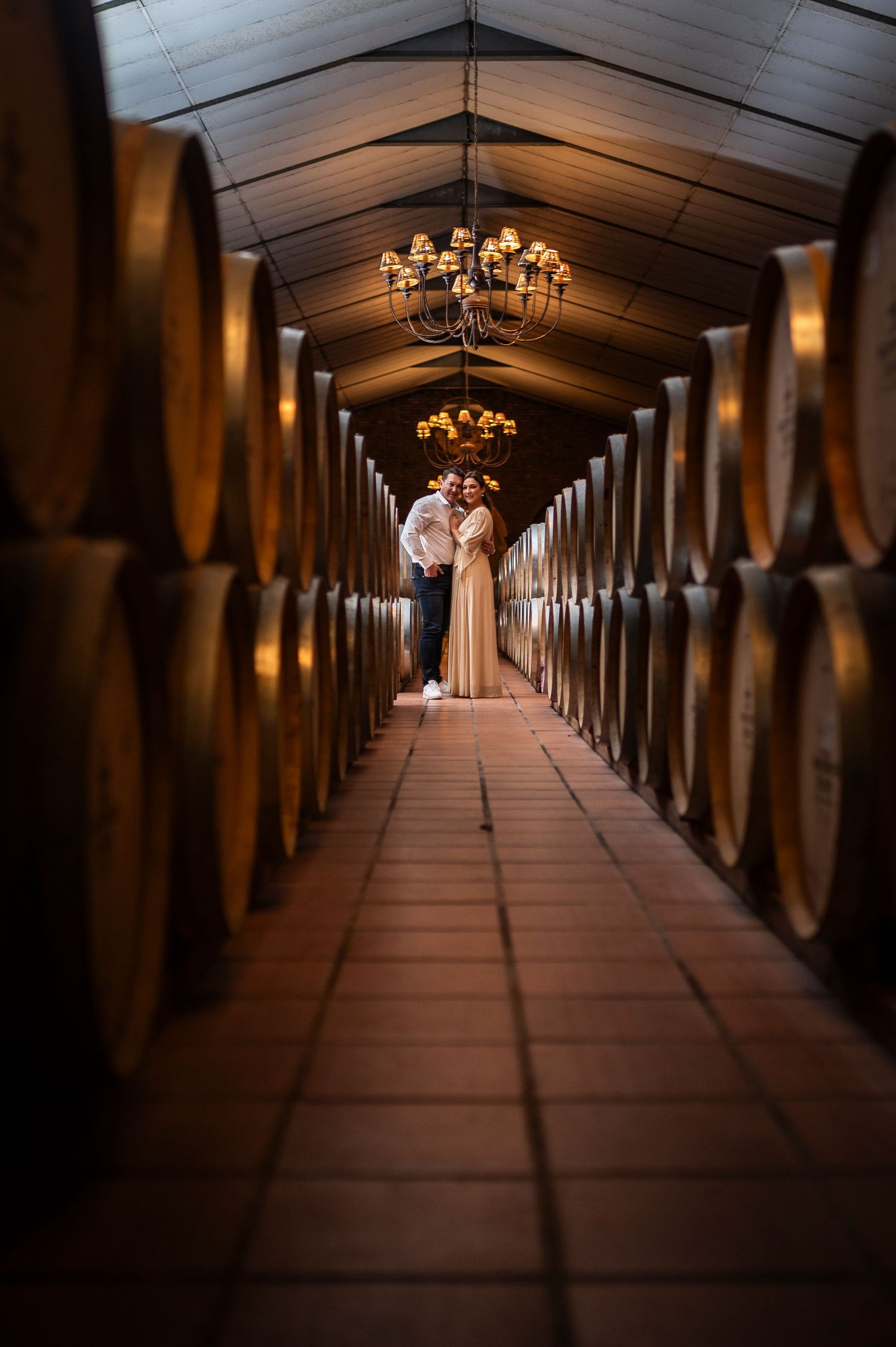 A couple stands silhouetted in a dramatic wine cellar tunnel lined with barrels and illuminated by elegant chandeliers.