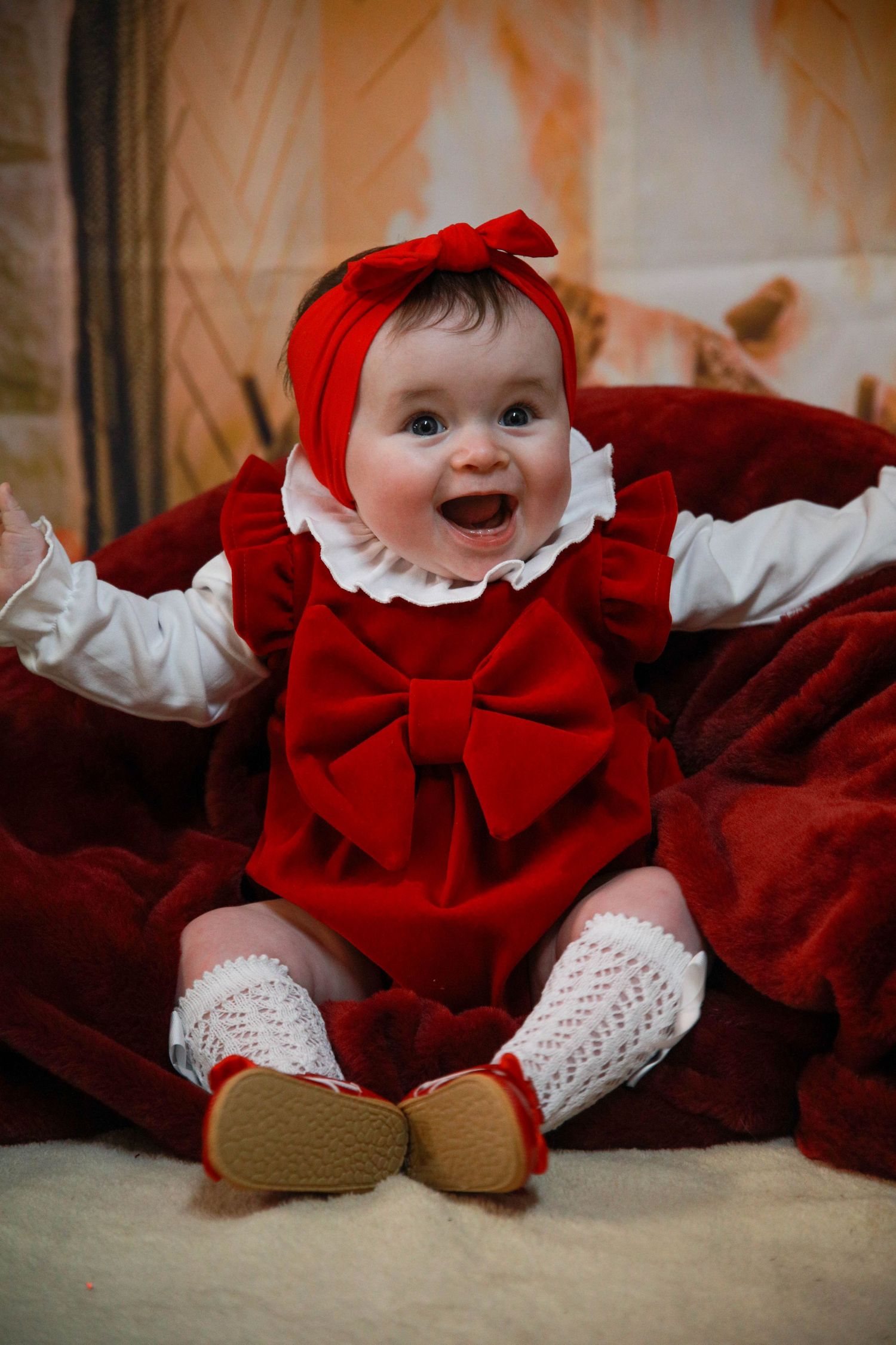 A smiling baby in a plaid holiday dress lies on a cream blanket with a snowman toy and winter decor in the background.
