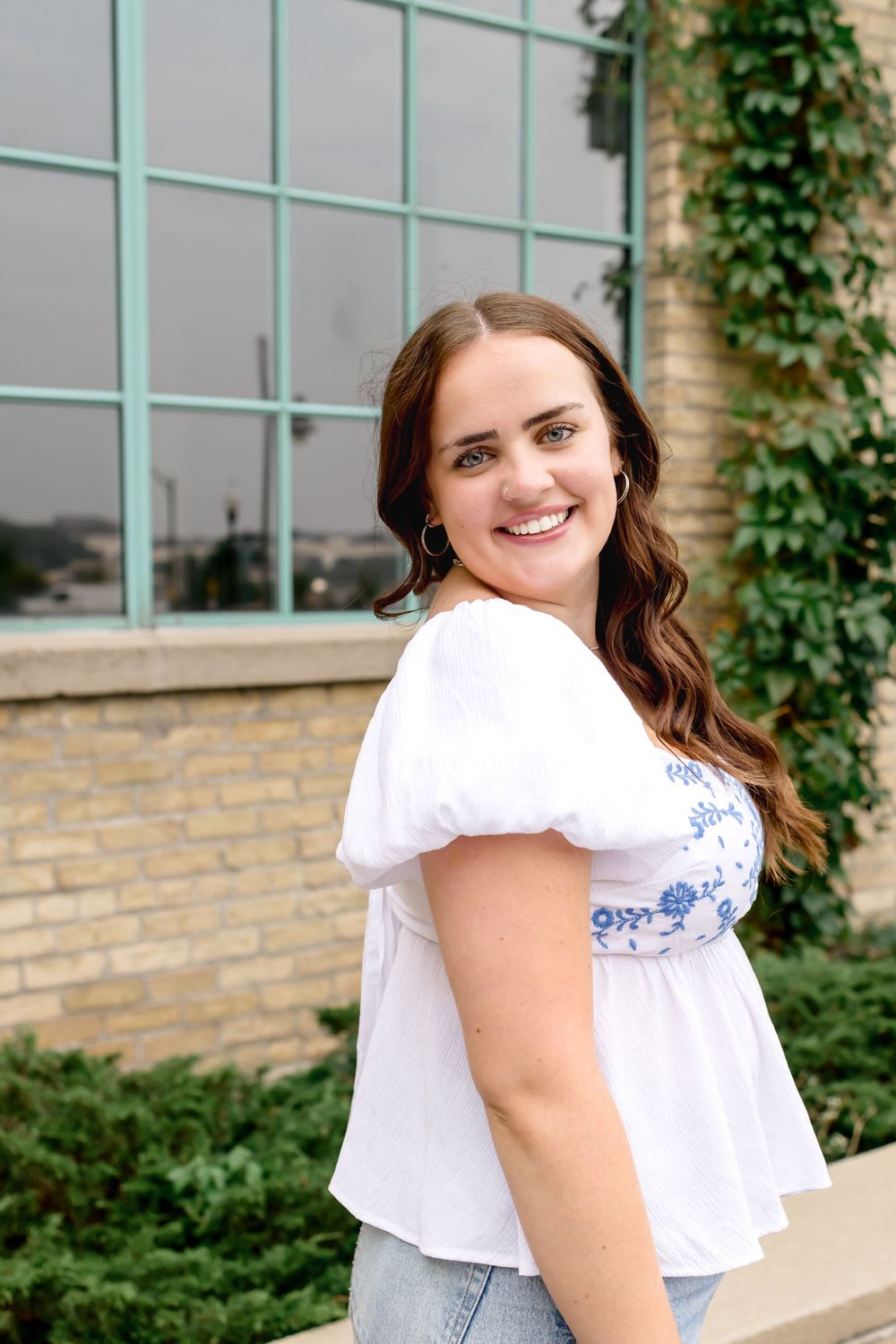 A woman in a white peasant top with blue embroidery poses in front of a brick building with climbing ivy.