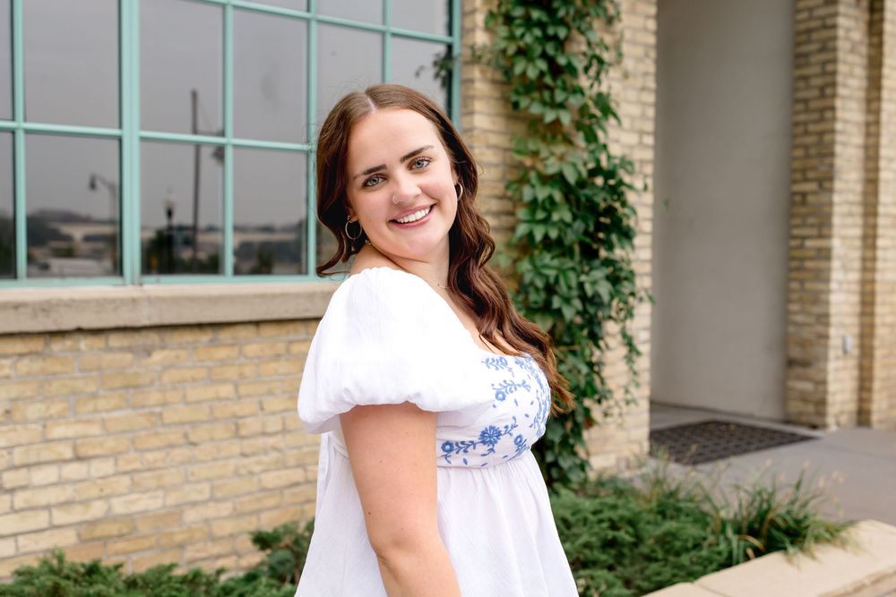 A woman wearing a white blouse with blue floral details smiles against a stone building exterior.