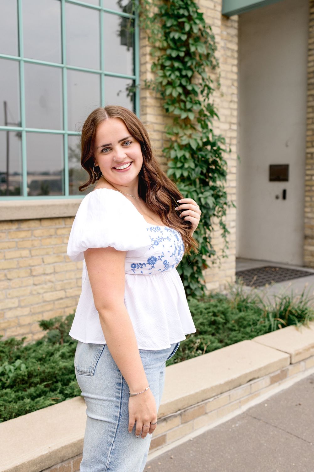 A casual outdoor portrait against a brick wall with ivy features a white puff-sleeve top and jeans outfit.