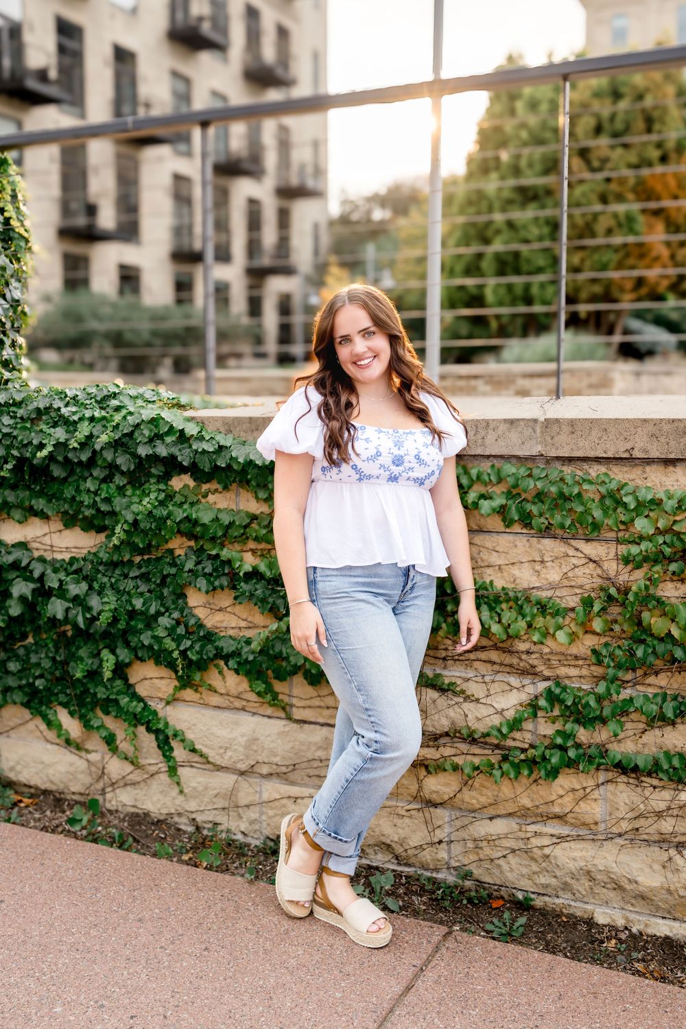 Golden hour portrait against ivy-covered wall showcasing summer casual wear with white top and jeans.