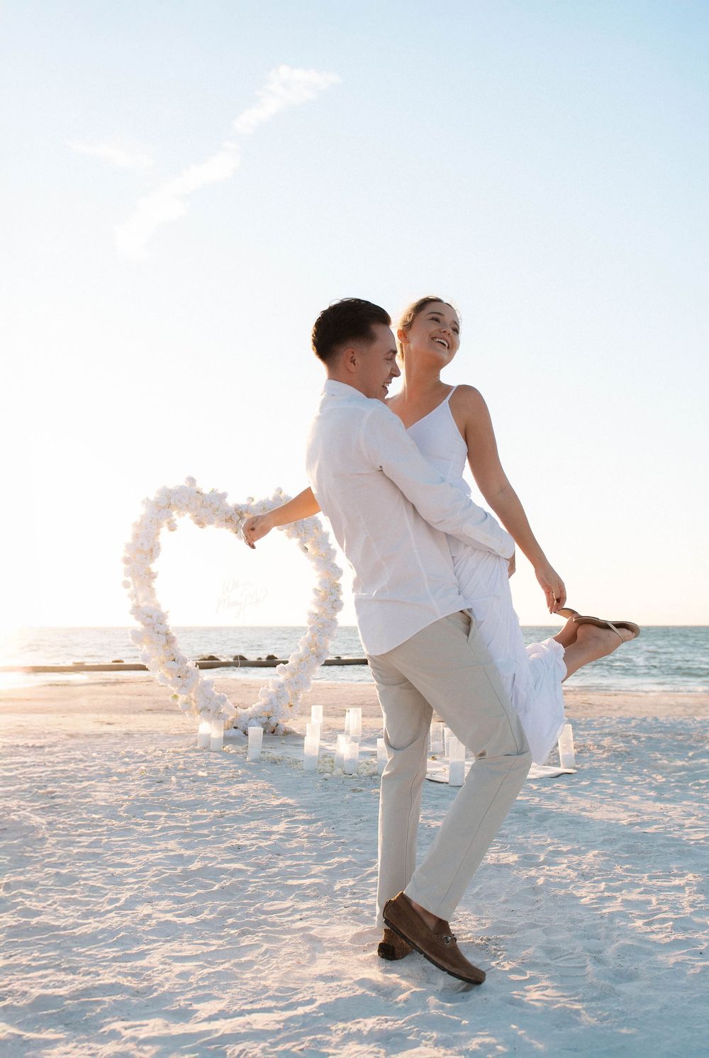 Couple celebrating their engagement beneath a white heart arch on Honeymoon Island, Florida.