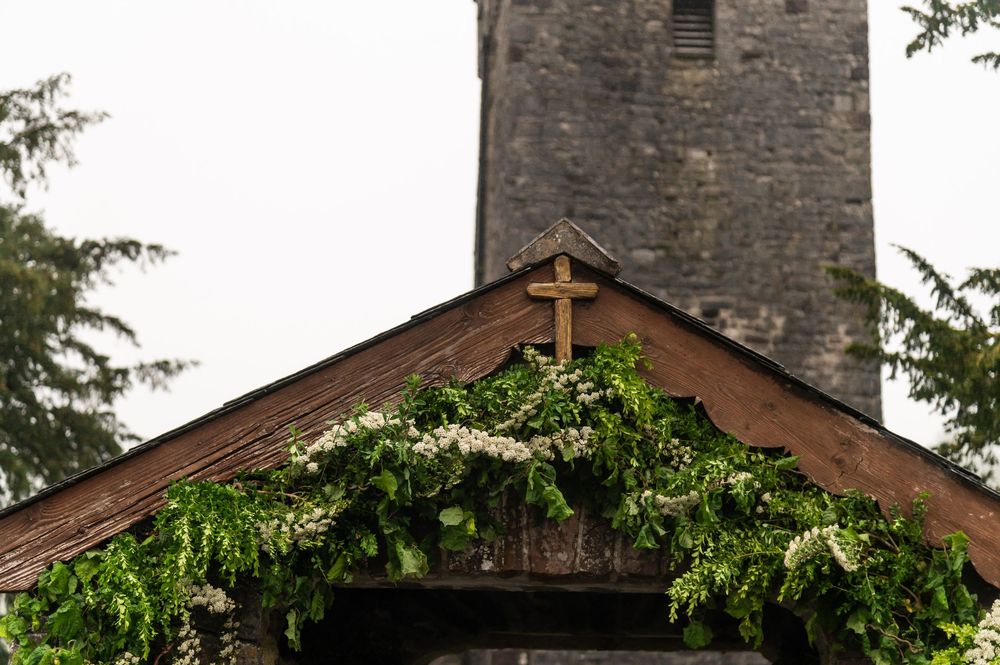Stone church with cross on top stands against cloudy sky surrounded by green trees.