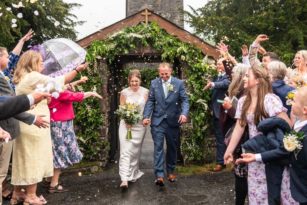 Wedding guests throw flower petals outside rustic stone church with greenery.