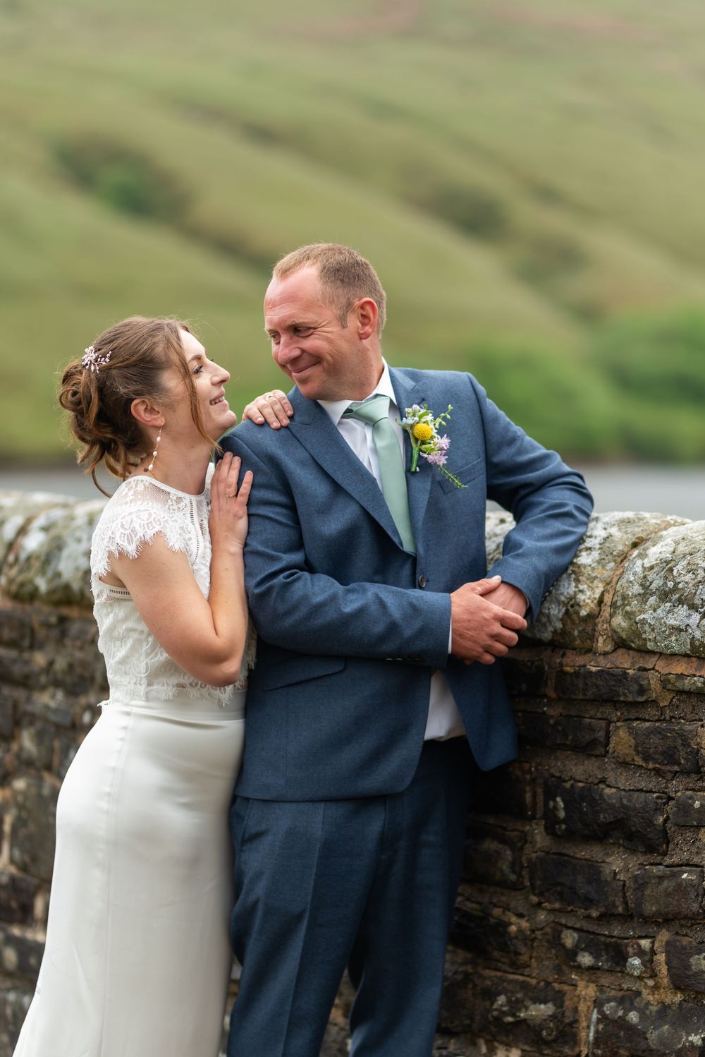 Couple shares intimate moment against rustic stone wall overlooking countryside.