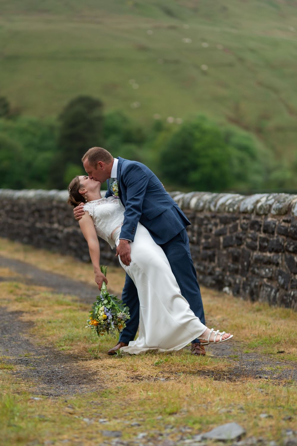 A romantic wedding photo of a couple in formal attire dipping against a scenic countryside stone wall backdrop.