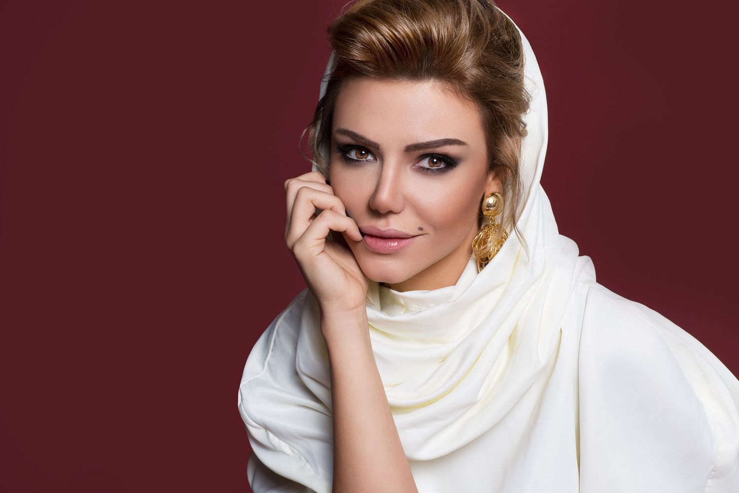 Portrait in a cozy white turtleneck sweater against a burgundy background with dramatic makeup and styled blonde hair.