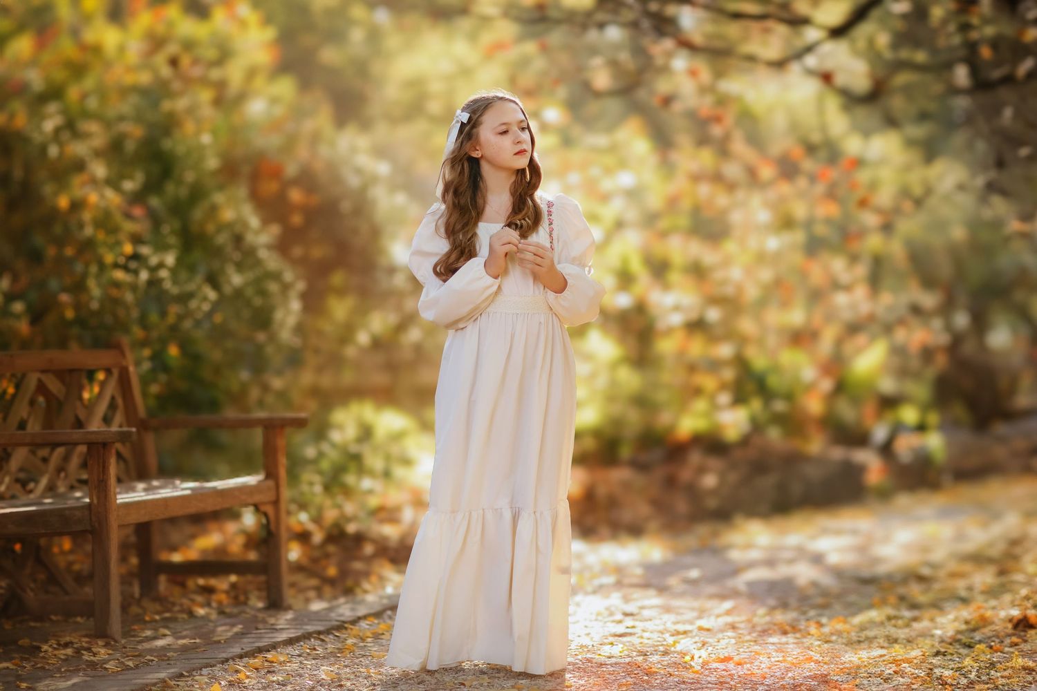 Woman in white flowy dress standing on path surrounded by autumn foliage in dreamy outdoor photoshoot setting.