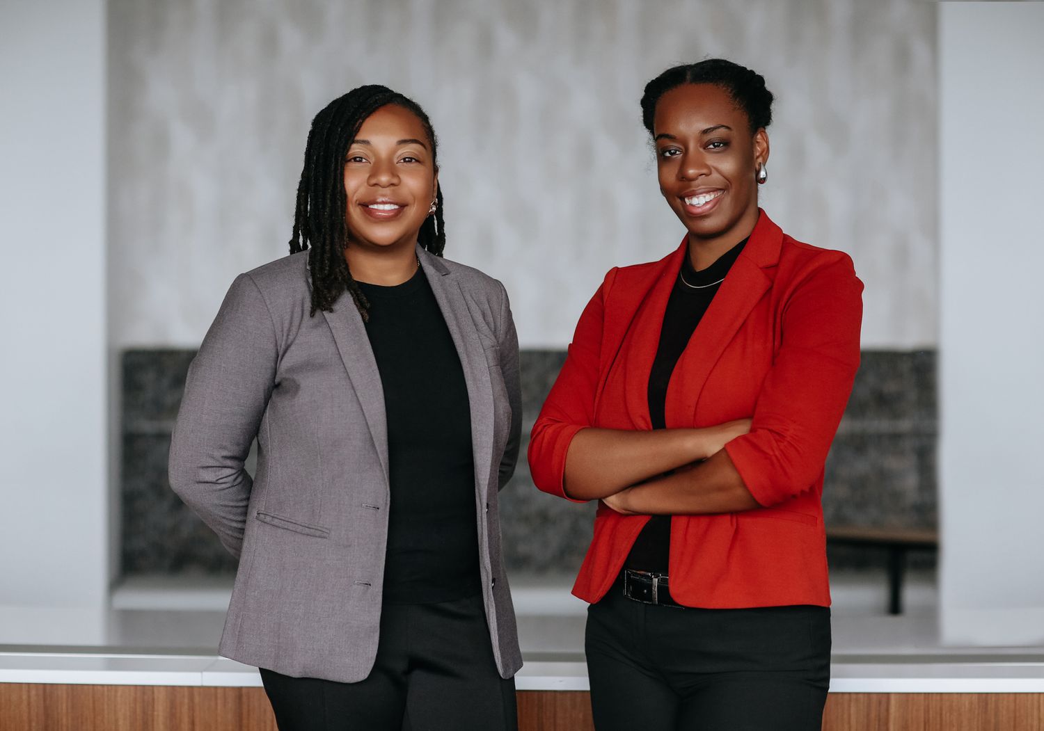 Two business professionals in blazers, one in gray and one in red, stand confidently against a light background.