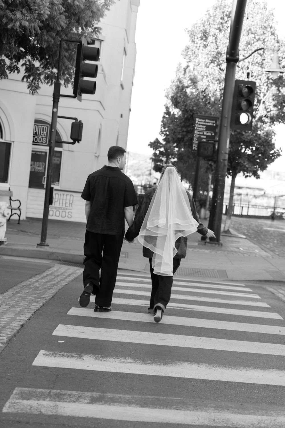 A couple walks hand in hand across a crosswalk in black and white, with one person wearing a flowing veil.