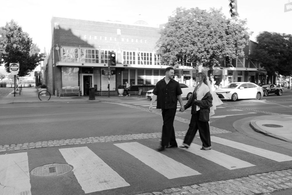 Two people share a moment while crossing a street in a black and white urban setting.