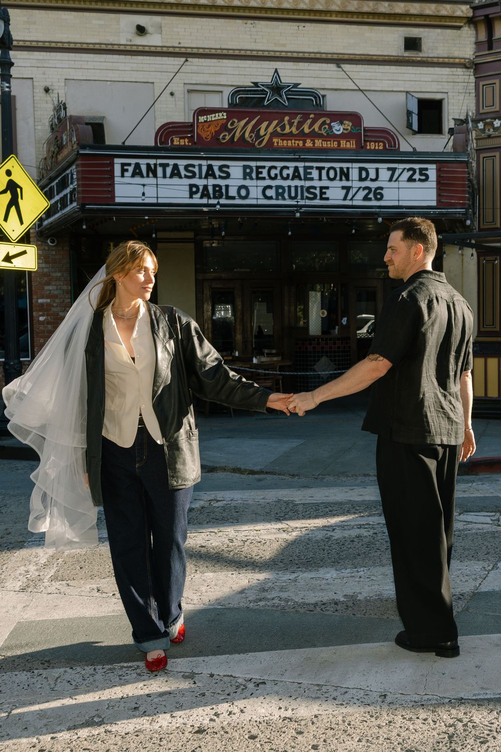 A couple in wedding attire poses outside a vintage theater marquee advertising reggaeton shows.