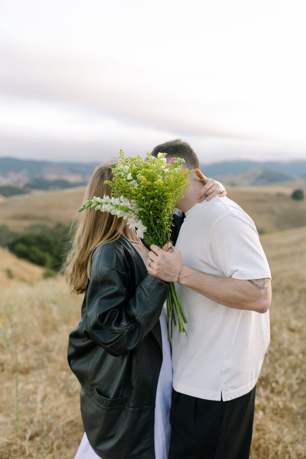 A couple embraces behind a bouquet of white wildflowers in a scenic mountain setting.
