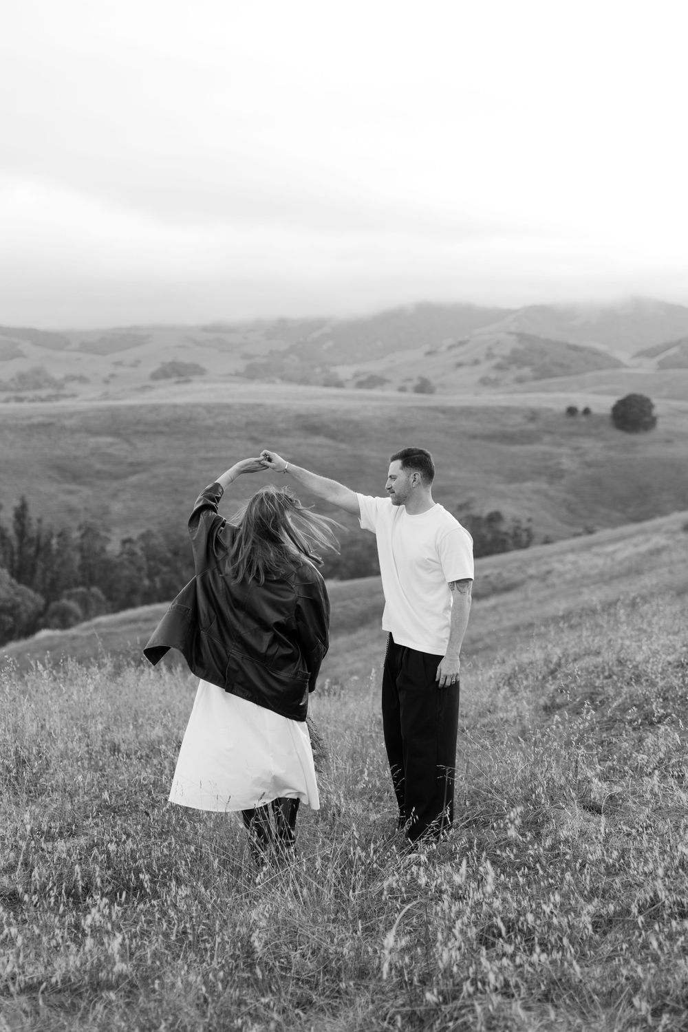 A couple shares a playful dance moment in a scenic hillside meadow.