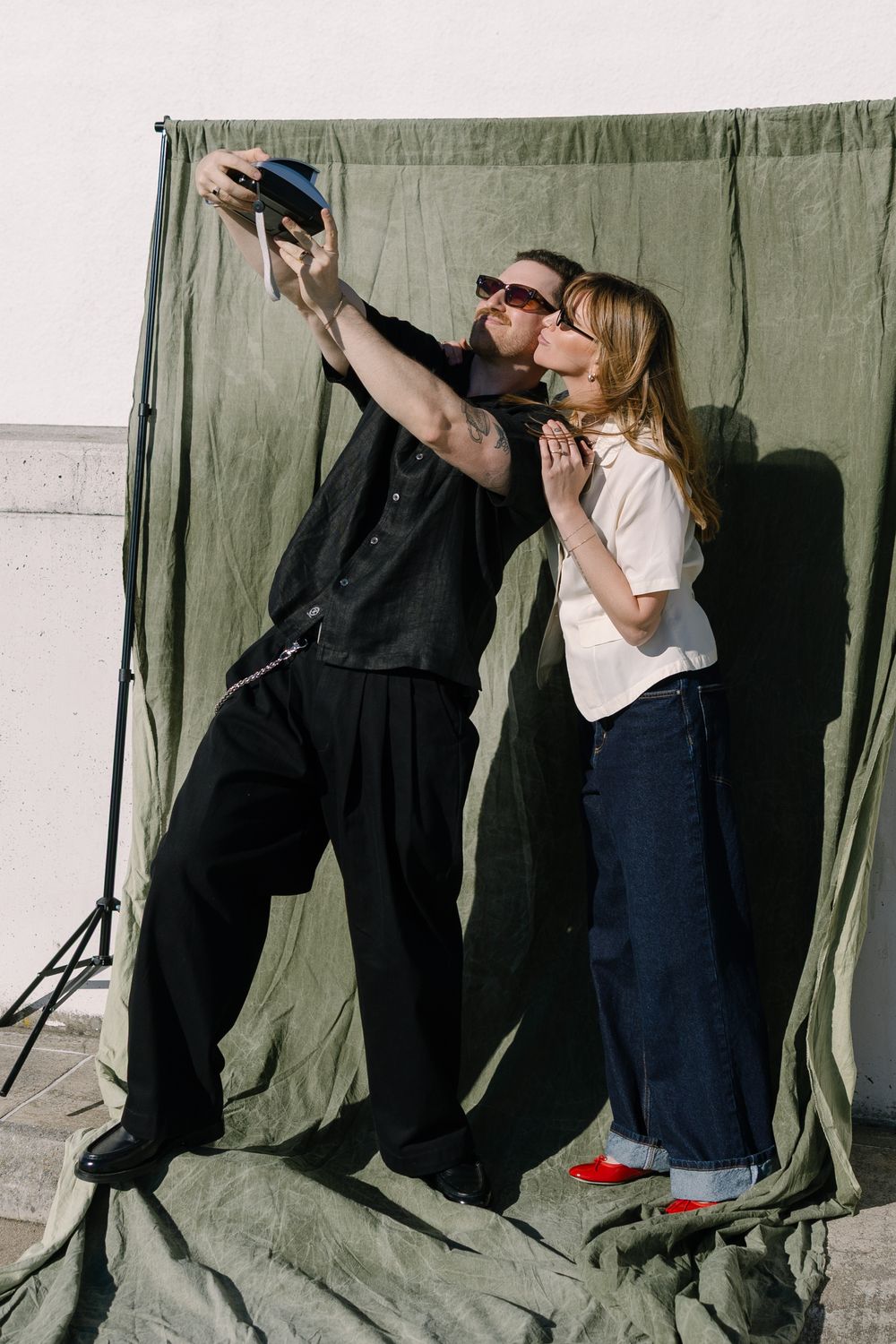 A playful couple takes a selfie against an olive green backdrop in casual attire.