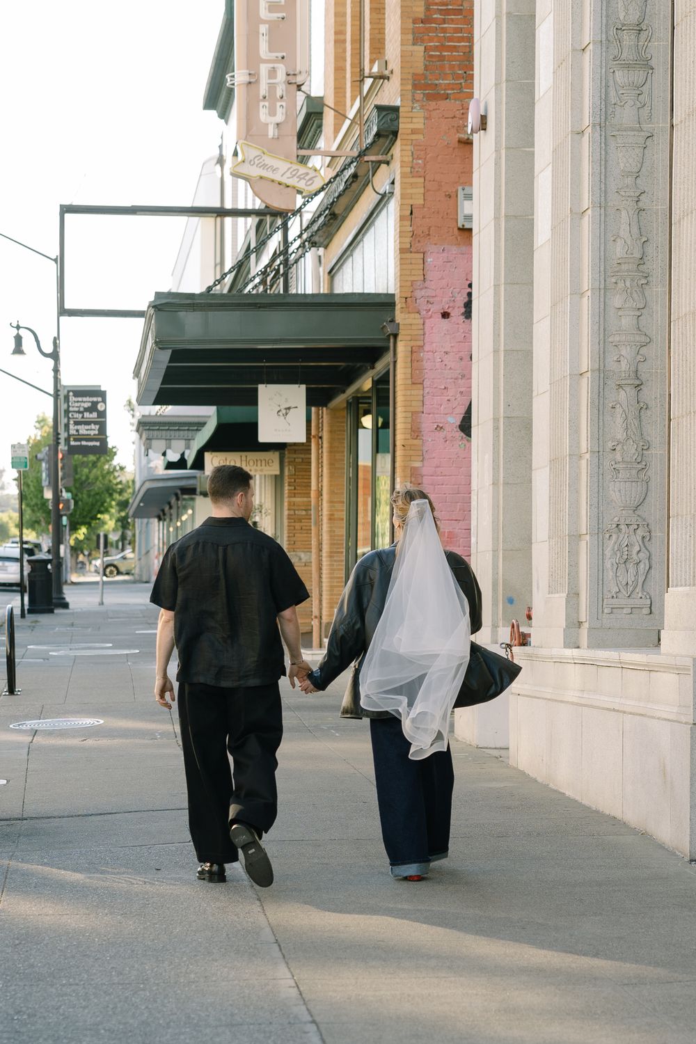 A couple walks hand in hand down a city sidewalk past historic storefronts.