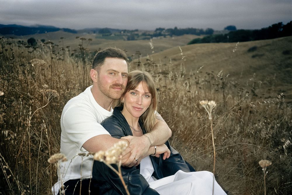 A couple embraces in a golden field of tall grass with mountains in the background.