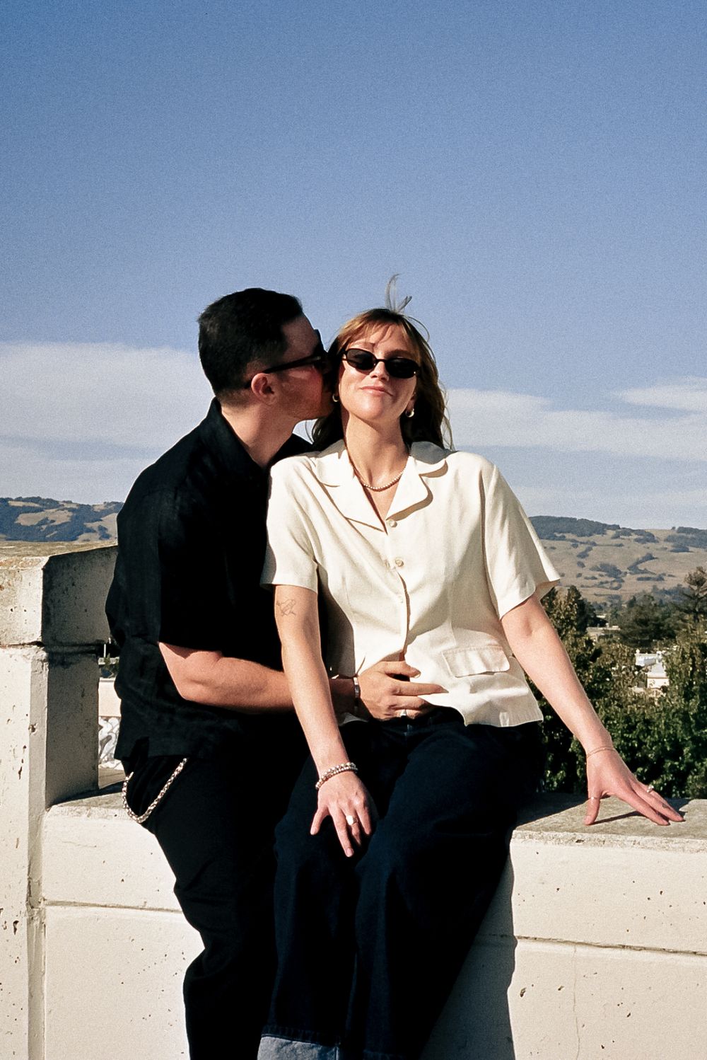 Two people in black and white clothing sit together on a white ledge against a bright blue sky.