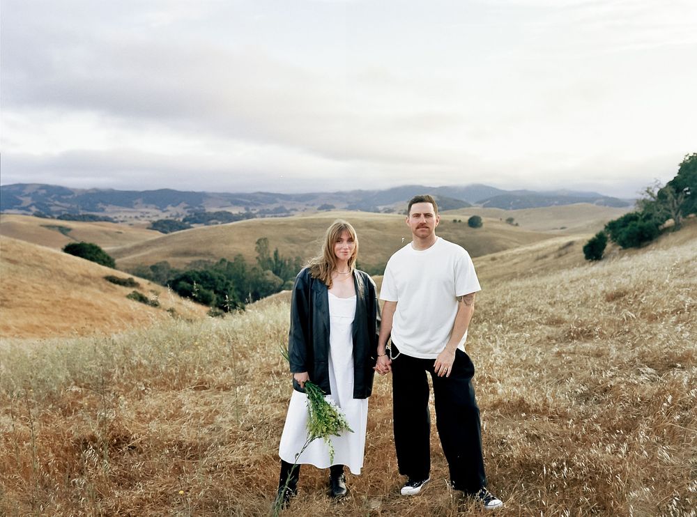 Two people stand together in a scenic field of dry grass with rolling hills in the distance.