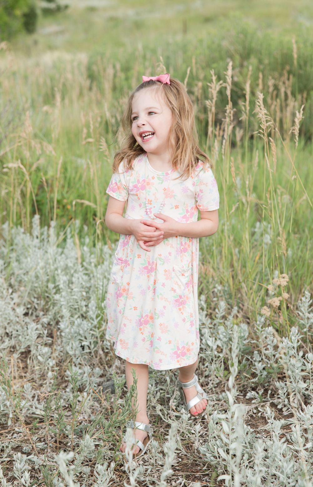 A joyful child in a floral dress laughs while standing in a meadow of tall grass and wildflowers on a sunny day.