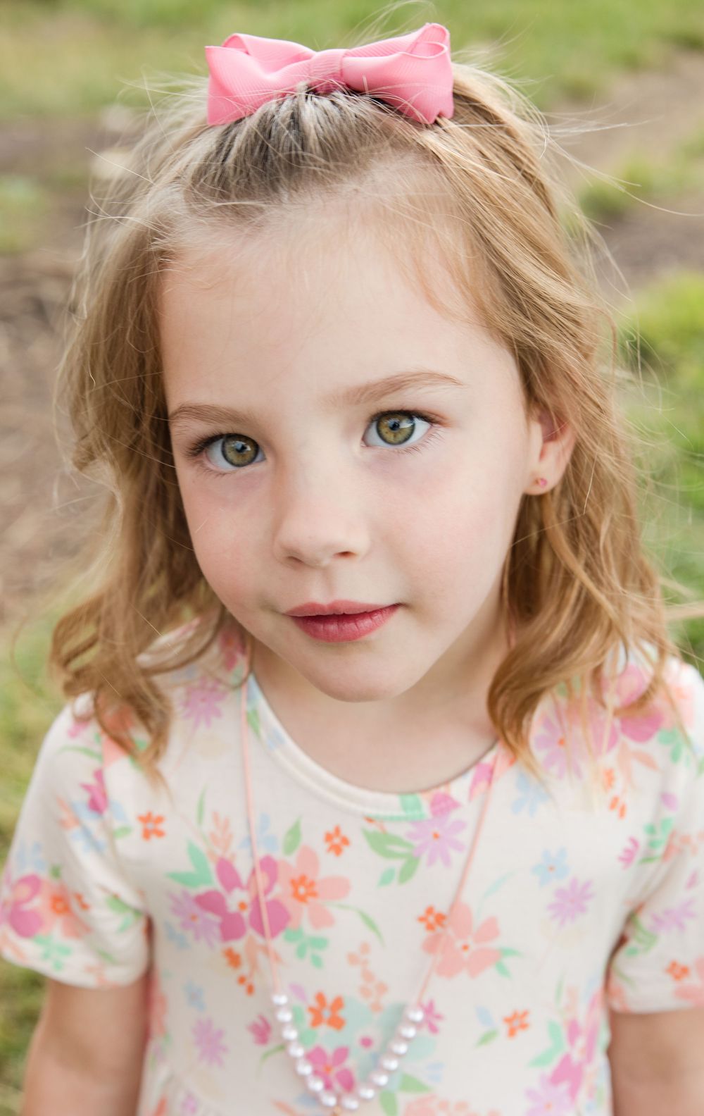 Sweet child wearing floral print dress and pink bow smiles at camera in outdoor portrait series.