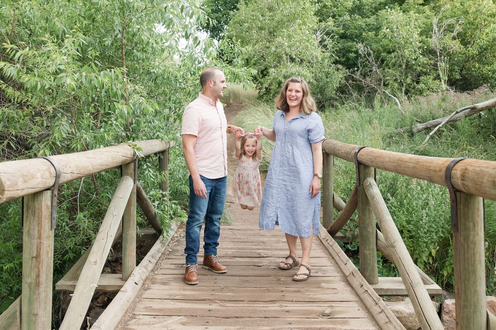 Two people and a small dog walk together on a wooden footbridge surrounded by lush green foliage on a sunny day.