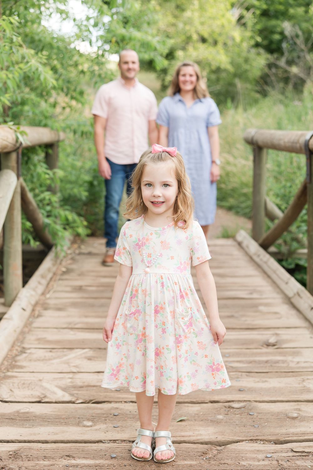 Family stands on a wooden bridge in a natural setting with trees and greenery in the background.