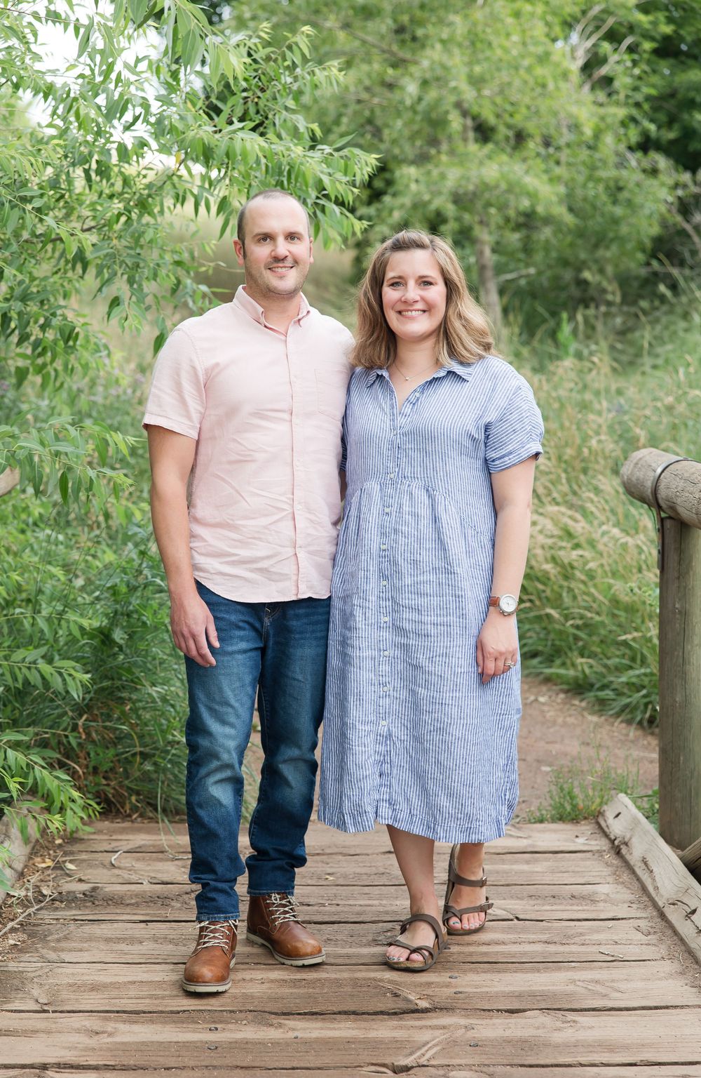 A couple stands smiling together on a wooden bridge in nature wearing casual summer clothing.