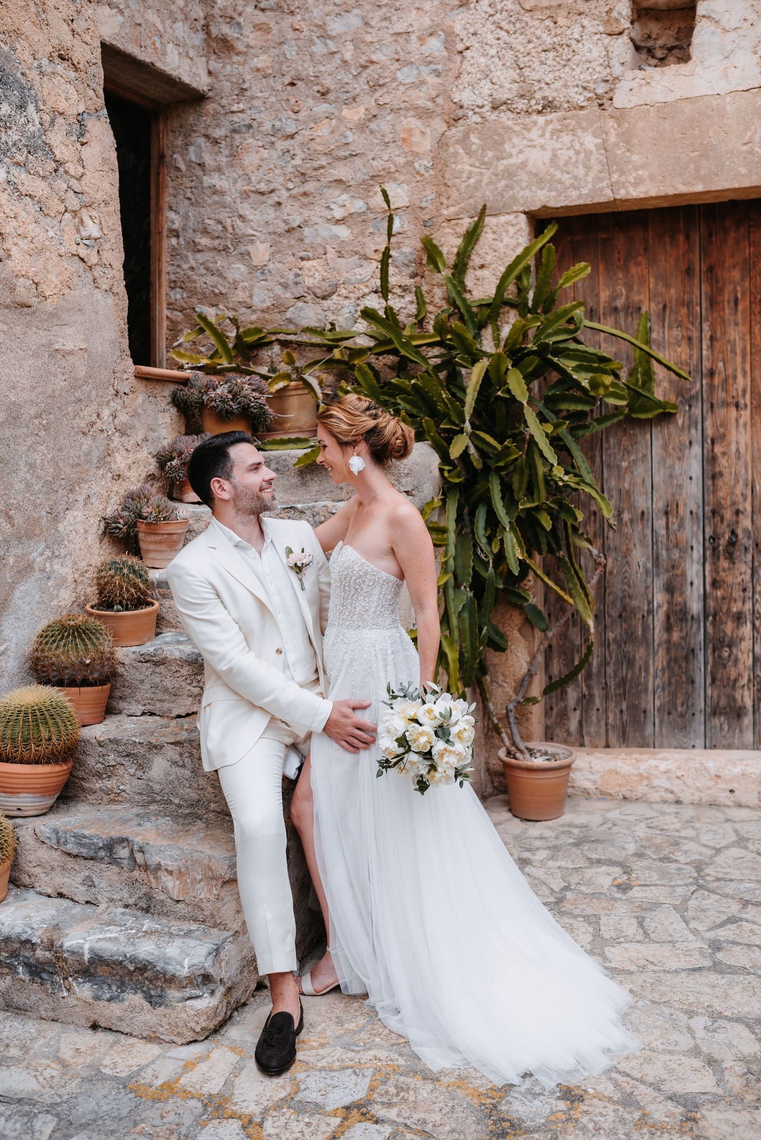 Ein Paar in eleganter weißer Hochzeitskleidung genießt einen zärtlichen Moment an einer rustikalen Steinmauer mit Topfpflanzen im Innenhof.