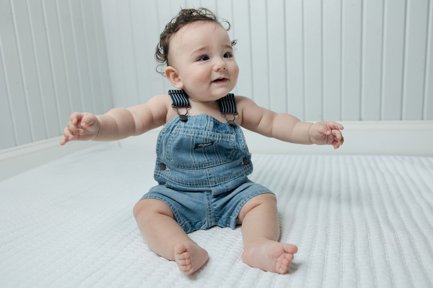 Cheerful baby in jean overalls sitting on white bedding with arms outstretched.