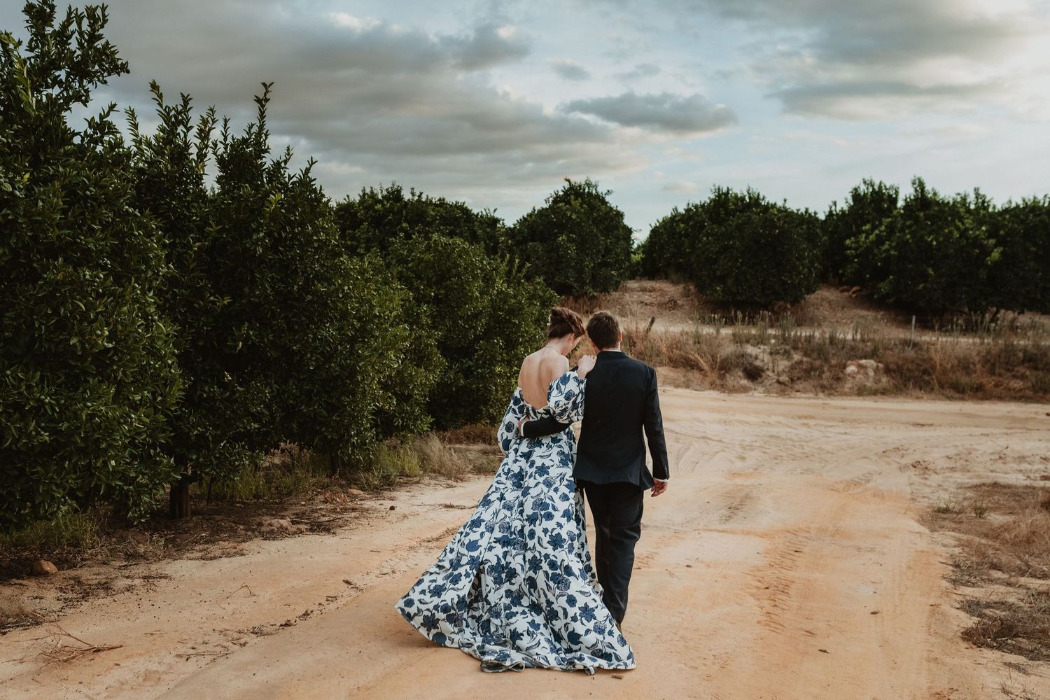 A couple walks hand in hand down a dirt path wearing formal attire surrounded by nature.