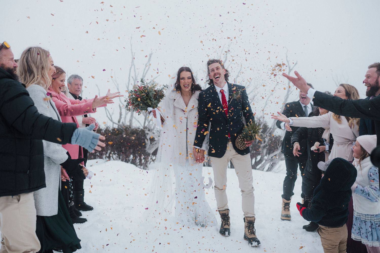 Cheering guests shower confetti and rose petals on winter wedding couple walking down a snowy path.