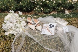 Wedding accessories including shoes, veil, and rings arranged on grass with white flowers in the background.