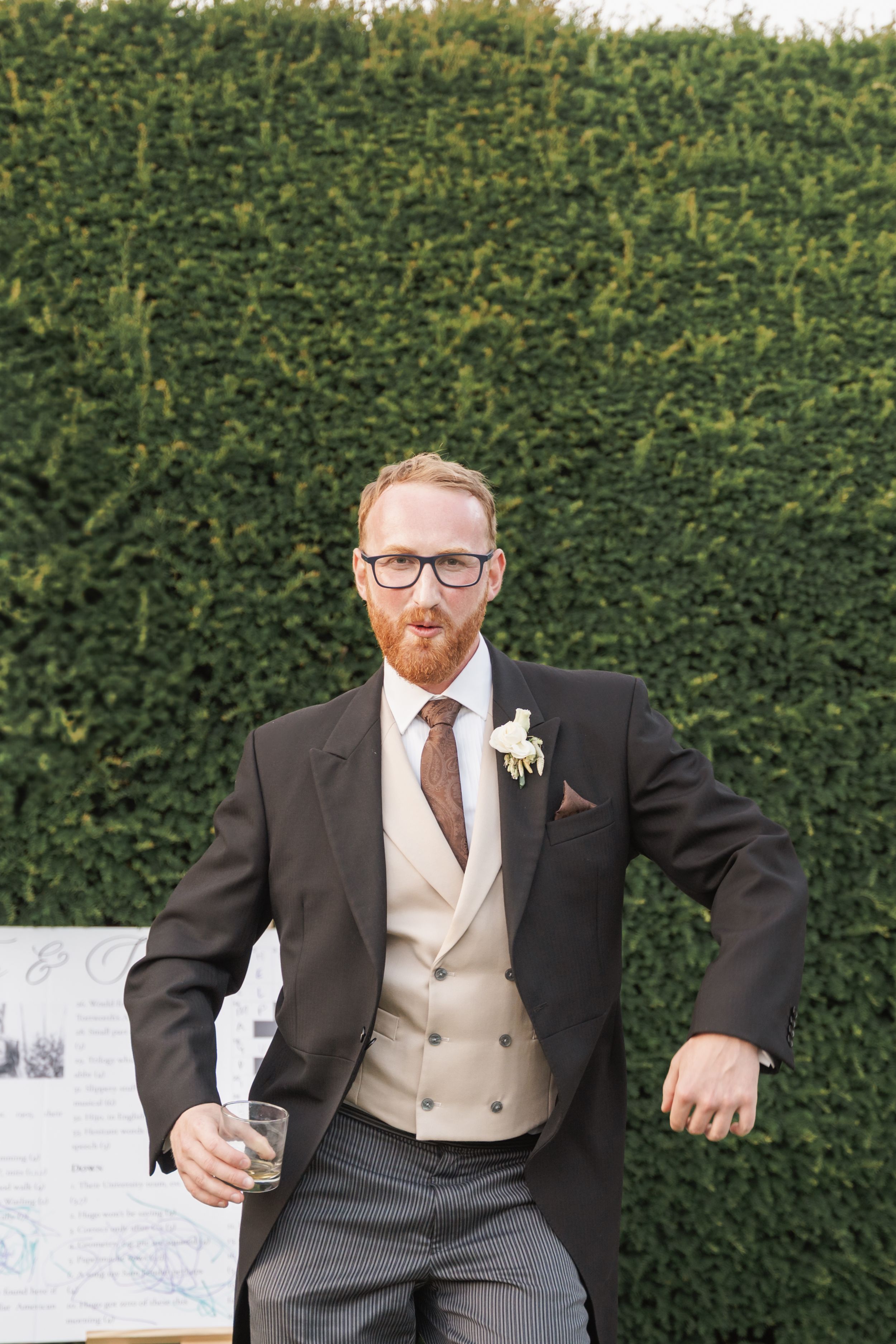 A person in a dark suit jacket, beige waistcoat and brown tie stands against a tall green hedge backdrop.