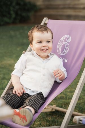 Happy toddler relaxes in a purple deck chair with number 6 design on a sunny lawn.