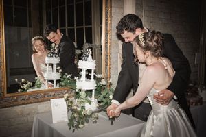 A couple cuts their elegant white tiered wedding cake decorated with greenery at their reception.