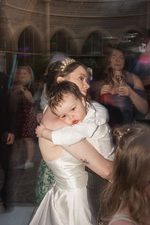 A tired child rests against a woman in a white wedding dress during a reception celebration.