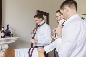 Three people in white dress shirts adjusting burgundy ties while getting ready in a boutique hotel room.
