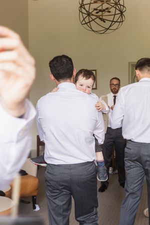 Groomsmen in white shirts and gray pants help each other get ready in a room with modern lighting.