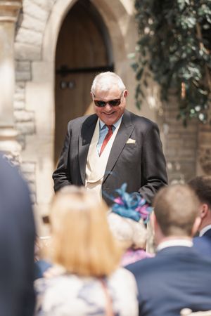 A formally dressed person in sunglasses and a suit smiles while standing among wedding guests outdoors.