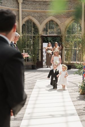 Wedding party members walk down an arched stone corridor decorated with greenery.
