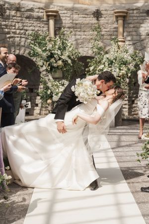 Wedding couple performs romantic dip pose against stone wall backdrop with floral decorations.